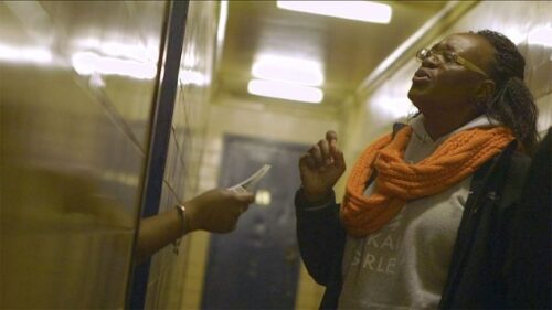 A black woman in a dim hallway wearing a Justice for Akai Gurley hoodie speaks to an unseen person behind an apartment door; the unseen person's hand is in frame holding a flier