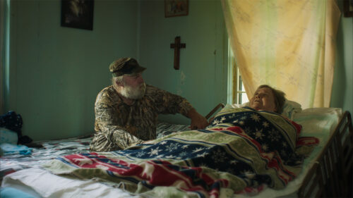 Man sitting next to a woman's bedside with a cross on the wall