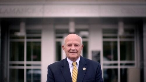 Sheriff Terry Johnson, an older bald white man in a business suit with a small badge on his lapel, stands outside a government building