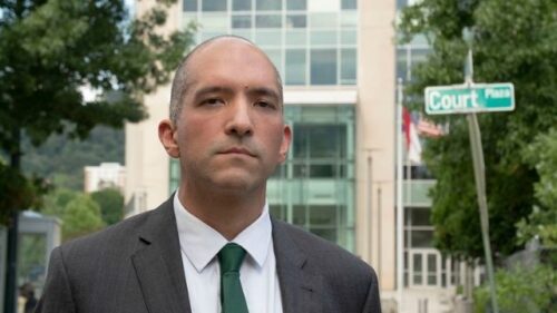 Public defender Ehsan Akhavi, a light skinned young bald man in a business suit outside a building with a road sign behind him that reads 
