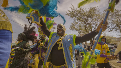 People in vibrant, colorful costumes dance and celebrate outdoors during a festive parade, with feathers, masks, and joyful expressions under a bright sky.