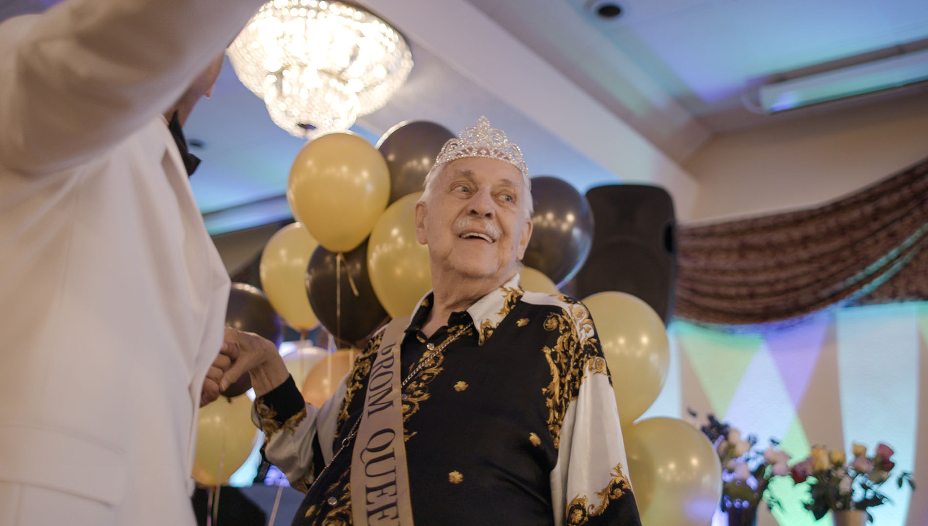 An elderly gentleman celebrates "prom" while in an assisted living facility, wearing sash and crown