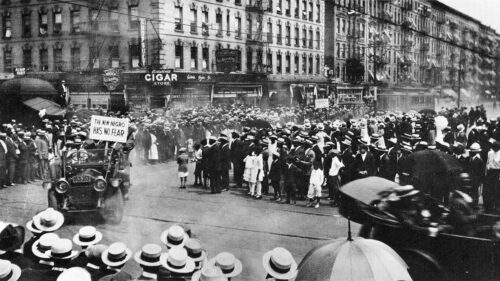Black and white old photograph of many black people standing at a street intersectionwith old car driving by with sign saying 