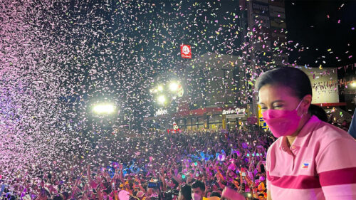 woman wearing pink shaking hands with a large crowd from a stage