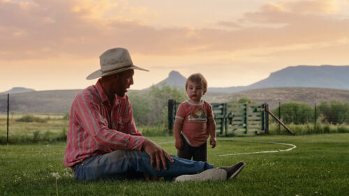 A man in a cowboy hat and a toddler stand in a grassy field with mountains under a sunset sky.