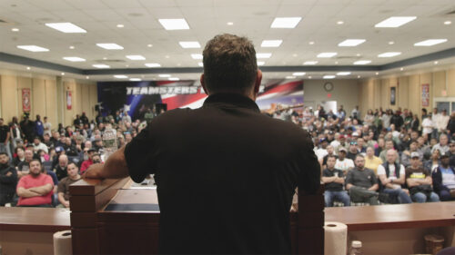 Man talking to a large group from a stage with a large poster that says 'teamsters' in the back of the room