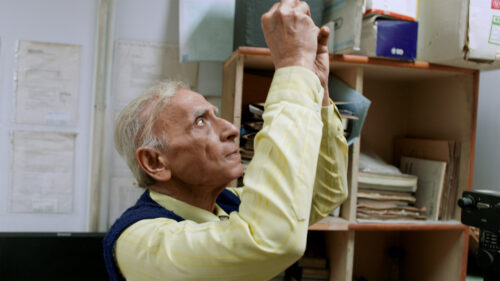 An older man in a yellow button-down shirt and blue sweater vest reaches toward a shelf filled with documents and boxes.