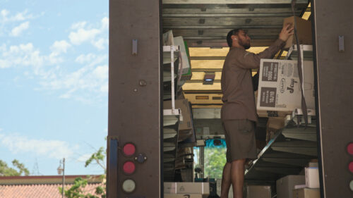 A delivery worker in a brown uniform places a cardboard box onto a metal shelf inside the back of a delivery truck.
