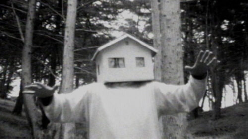 black and white photo of man in woods with model house on his head