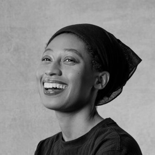 A woman wearing a headscarf smiles broadly, looking slightly to the side. The photo is black and white with a plain, textured background.