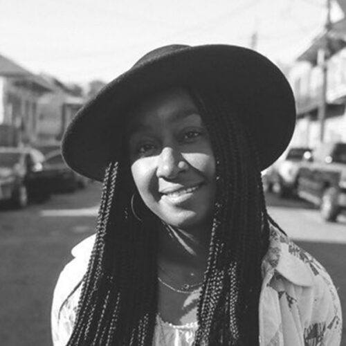 A woman with long braids and a wide-brimmed hat smiles outdoors on a street lined with parked cars, captured in black and white.