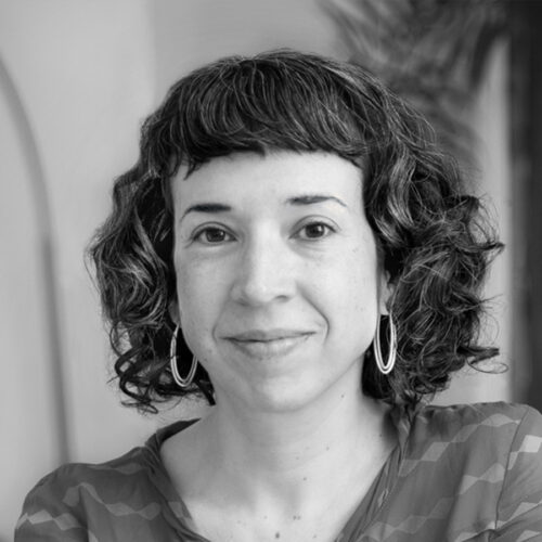 A woman with short, curly hair and hoop earrings smiles softly at the camera. She is wearing a patterned top. The photo is in black and white.