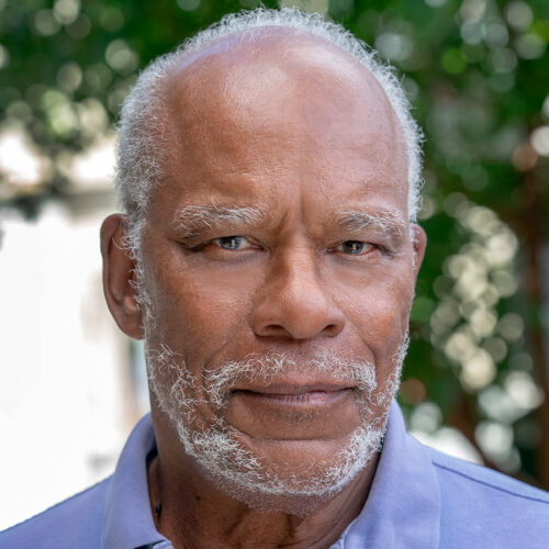 Older man with short white hair and a beard, in a light blue collared shirt, gazes seriously at the camera. Soft-focus green foliage fills the background.
