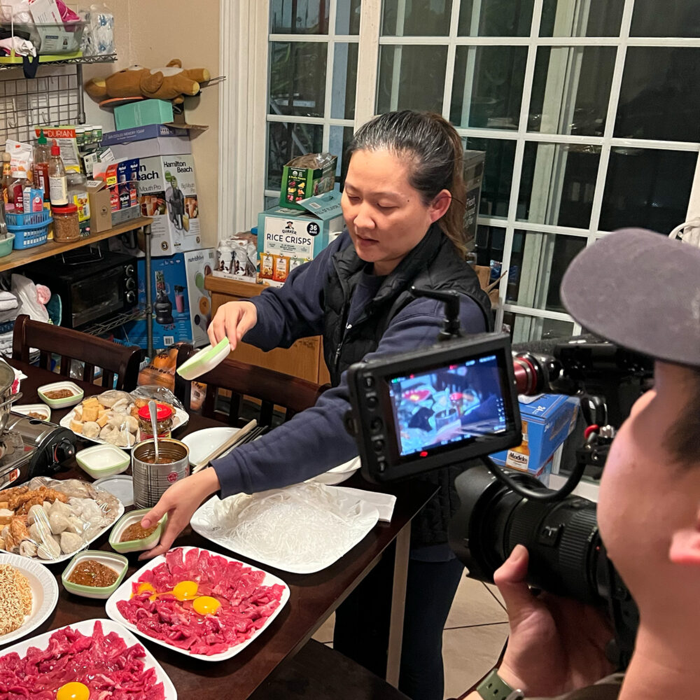 Person holding camera shooting a woman with dark brown hair and medium tan skin tone who is preparing food