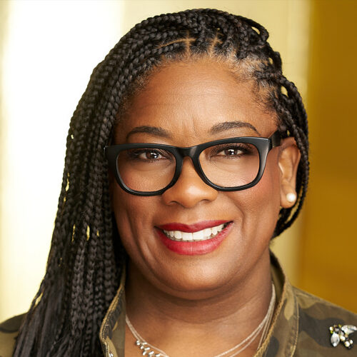 A woman with long braided hair, black glasses, red lipstick, a camouflage jacket, and layered necklaces stands smiling in front of a golden-yellow background.