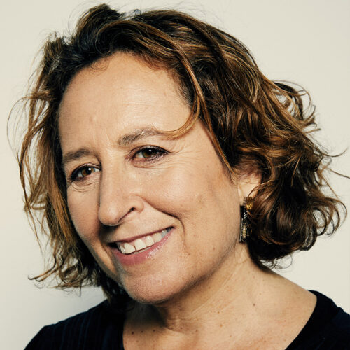 Smiling woman with short, wavy brown hair, wearing a dark top and gold earrings, in front of a plain light background.
