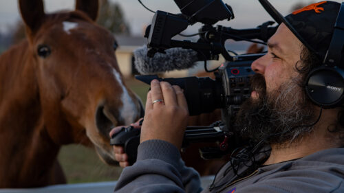 Man looking through camera outside with horse in background looking at man