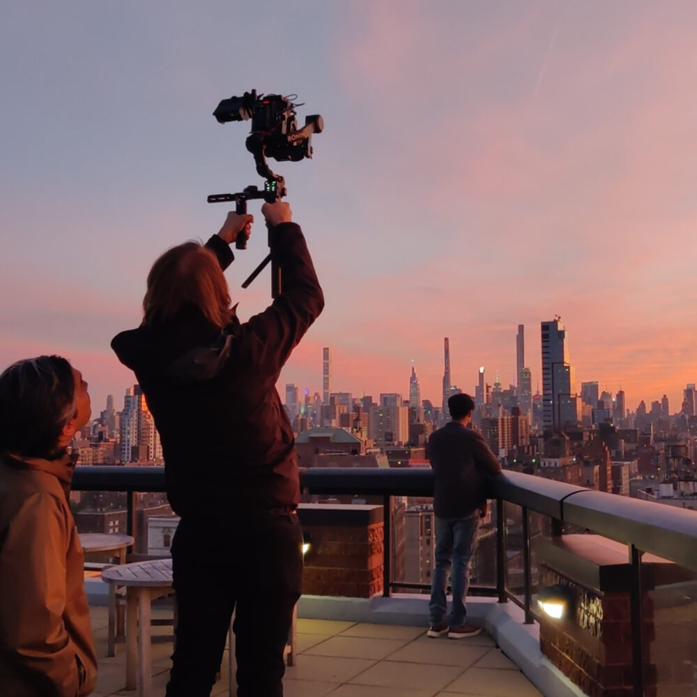 Three people on a rooftop at sunset looking out over the New York skyline. Camera looking at man