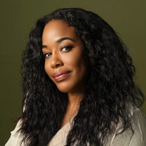 A woman with long, curly black hair and a light beige top smiles softly against an olive green background.
