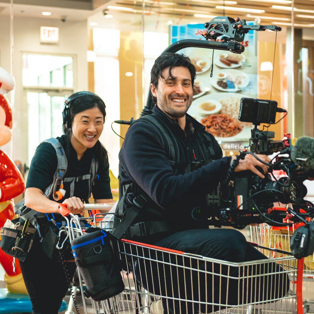 Man holding a camera while sitting in a shopping cart with a woman also wearing camera gear both smiling