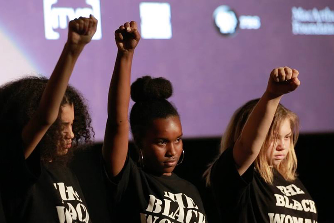 Three women holding their fists in the air looking out of frame and wearing shirts that say The Black Women. Serious expressions on their face
