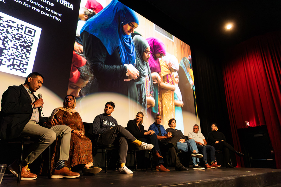 A divers group of people on a stage sitting in a row. one person holding a mic. Film publicity still on a screen behind them.