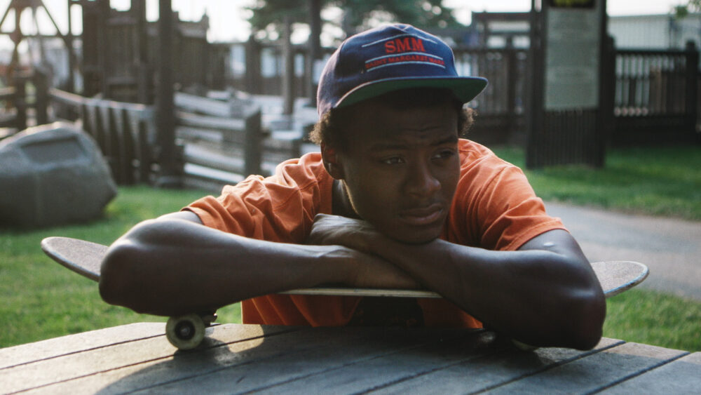 Young man resting his arms and head on a skateboard wearing a hat with dark brown skin tone looking into the distance