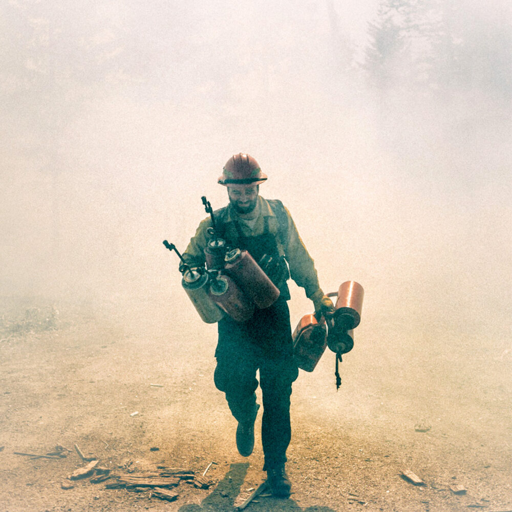 Person wearing fire gear walking toward the viewer carrying various things. Very smokey air around them.