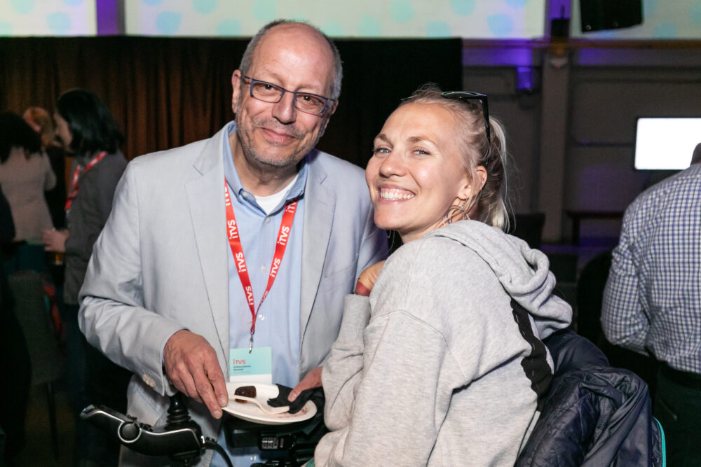 ITVS employee who is a light skinned older man with receding grey hair and glasses wearing a suit and employee badge smiling with apparent filmmaker who is light tan skin tone with light blond hair in a mobility chair.