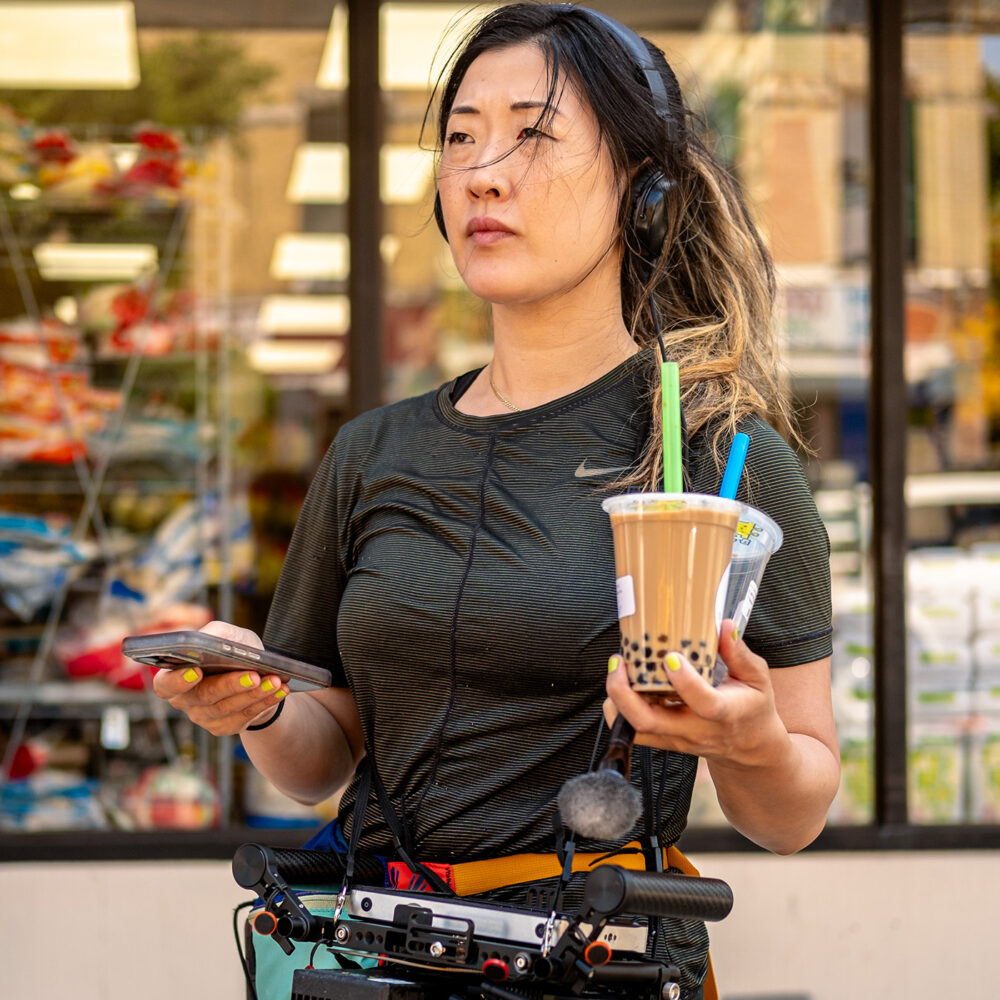 Woman with dark hair and fair complexion wearing headsphones and camera gear around waist holding phone in one hand and two bobas in the other
