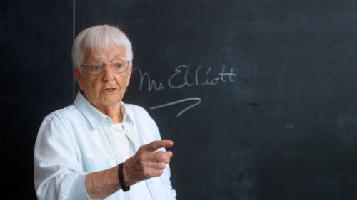 An elderly person with short white hair and glasses stands in front of a chalkboard, pointing forward. Mrs. Elliott is written in cursive on the board behind them.