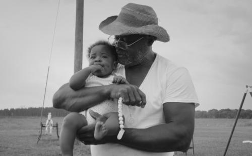 Man holding a baby in a field kissing the top of their head. Both with dark skin tones and dark eyes