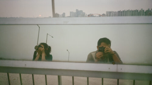 Two people are reflected in a glass panel, each holding a camera and taking a photo. A cityscape and water are visible in the background.