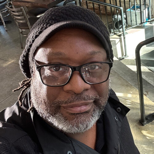 A person with glasses, a black knit cap, and a salt-and-pepper beard taking a selfie indoors. Sunlight shines on their face, and there are metal railings and chairs in the background.