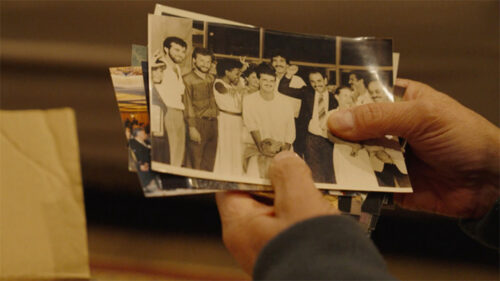 A person holds a stack of photographs, with the top photo showing a group of people posing and smiling indoors.