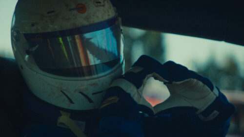 A race car driver wearing a helmet and gloves sits in a car, forming a heart shape with their hands.