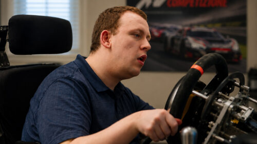 A person in a blue shirt sits in a chair holding a racing simulator wheel, with a poster of race cars on the wall in the background.