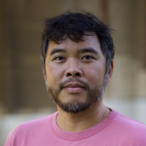 Man with short dark hair and beard, wearing a pink shirt, stands outdoors facing the camera; background is blurred in brown and light tones.