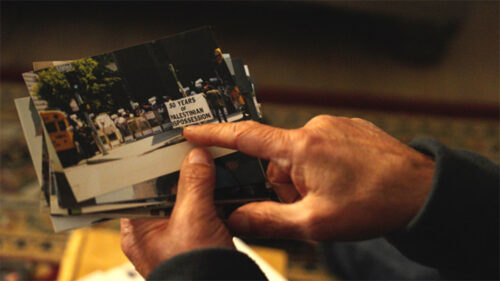 A person holds a stack of photographs, with the top photo showing a group of people posing and smiling indoors.