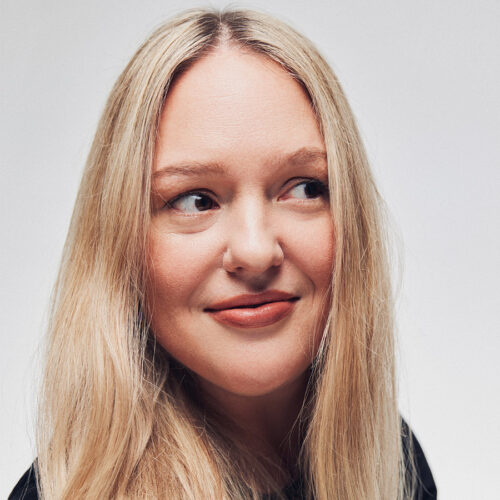 A woman with long blonde hair and light skin looks slightly to the side, smiling softly, against a plain light background.