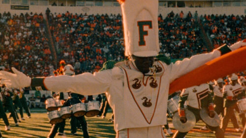 Marching band member in a tall white hat with an F, white uniform, and green accents performs on a football field, with drumline and a crowd in the background.