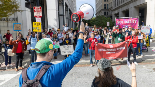 A crowd of protesters holds signs like DEFEND THE CONSTITUTION and SOLIDARITY while a person in a green cap raises a megaphone, speaking to the group on a city street.