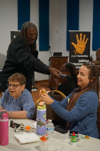 A man holds a microphone for a smiling woman at a table with snacks and drinks, while another woman sits beside them. Project Humanities banners are visible in the background.
