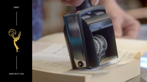 A close-up of a hand stamping a date on a piece of paper resting on an open book, with an Emmy nomination graphic on the left side.