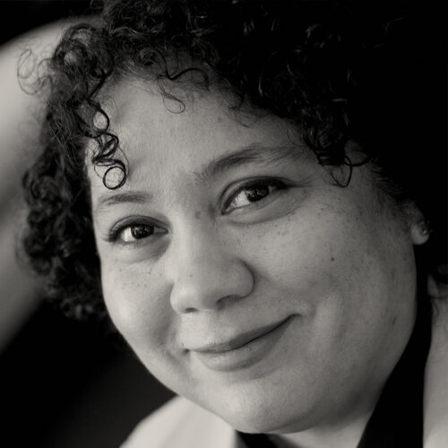A person with curly hair smiles softly while looking at the camera in this close-up, black-and-white portrait.