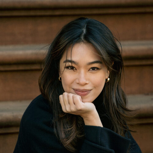 A woman with long dark hair and gold hoop earrings smiles softly, resting her chin on her hand, seated on brown steps outdoors.