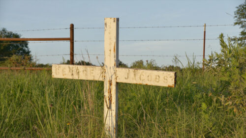 A weathered white wooden cross with faded text stands in tall grass near a barbed wire fence on a sunny day.