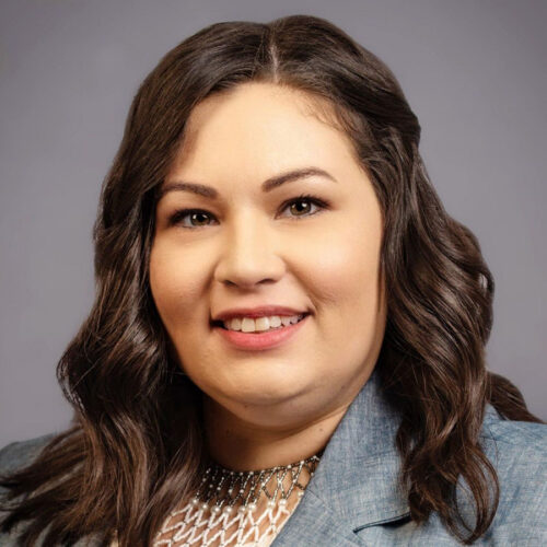 A woman with wavy dark brown hair, wearing a blue blazer and a decorative necklace, smiles at the camera against a plain gray background.