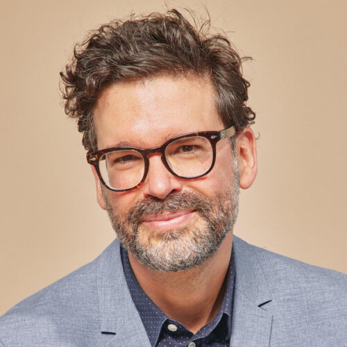Smiling man with curly brown hair, beard, and glasses wearing a light blue blazer and dark shirt against a beige background.