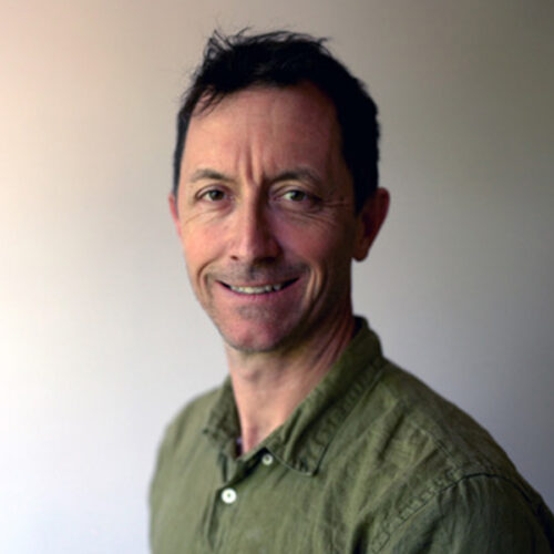 A man with short dark hair and a slight smile is wearing a green button-up shirt, standing in front of a plain, light-colored background.